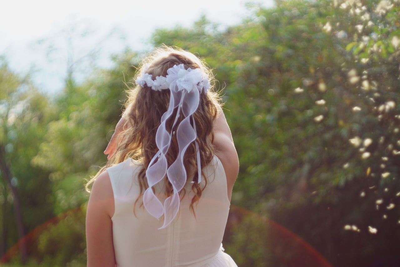 niña vestida de blanco y con un lazo en el pelo, de espaldas realizando su primera comunión